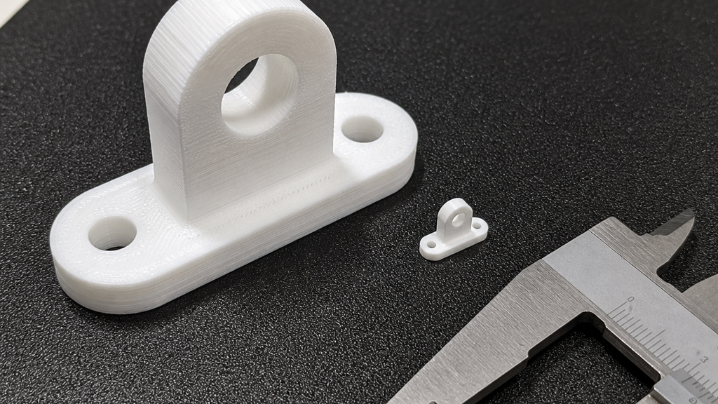 Large and miniature versions of the same white bracket beside calipers, illustrating a scale mismatch.